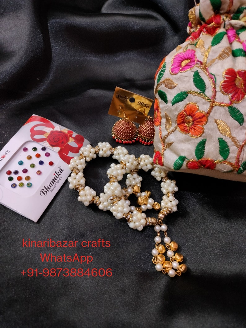 May include: A collection of jewellery and accessories displayed on a black satin background. The image features a white beaded necklace with gold details, red and gold earrings, a floral embroidered pouch, and a sheet of colourful bindi stickers.