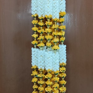 May include: A long garland of yellow and white artificial flowers with gold bells at the bottom. The garland is made of fabric flowers and is perfect for decorating a home or event.