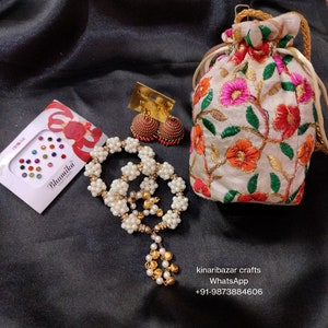 May include: A collection of jewellery and accessories displayed on a black satin cloth. The image shows a white beaded necklace with gold details, red and gold earrings, an embroidered pouch, and face gems.