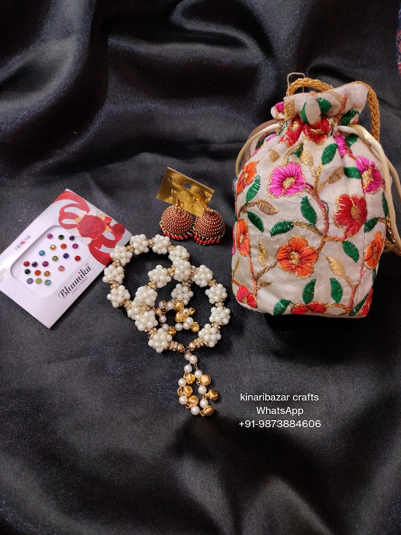 May include: A collection of jewellery and accessories displayed on a black satin cloth. The image shows a white beaded necklace with gold details, red and gold earrings, an embroidered pouch, and face gems.