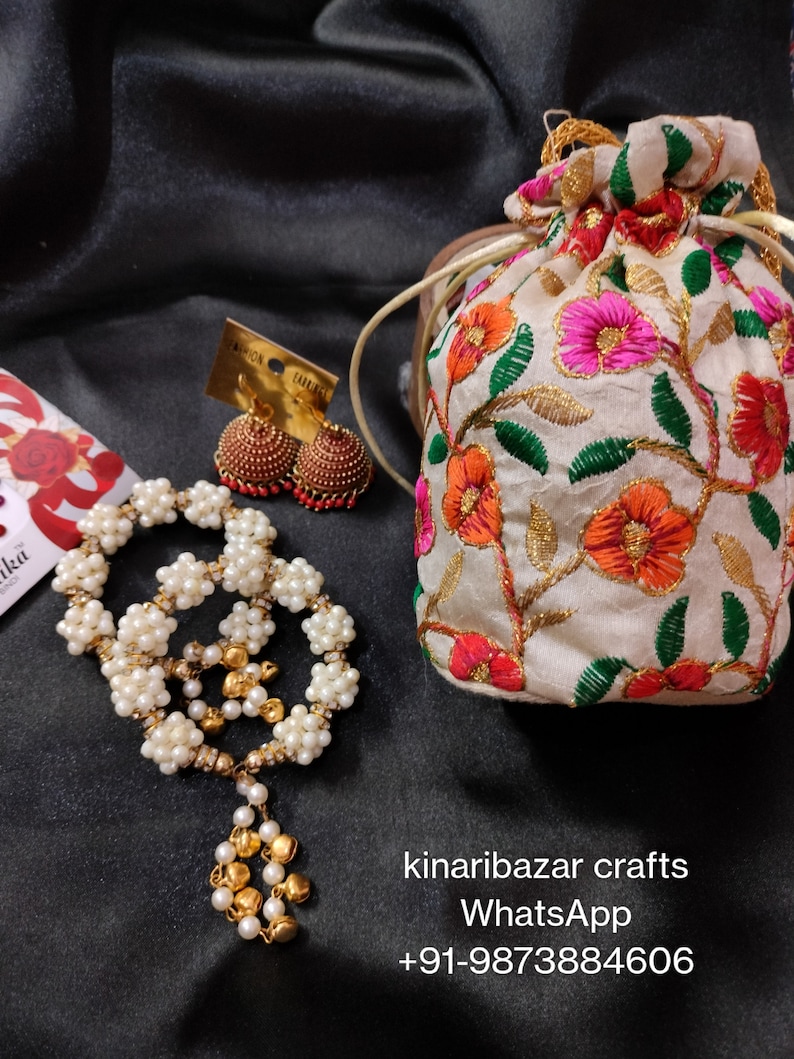 May include: A collection of handcrafted jewellery and accessories. Features a white beaded necklace with a gold pendant, gold and red earrings, and a floral embroidered pouch. The image also displays the text "kinaribazar crafts" and a WhatsApp number.