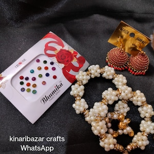May include: A collection of Indian jewellery and accessories. The image shows a box of colourful bindi stickers, gold-toned earrings with red detailing, and a beaded necklace with pearl-like beads and gold accents. The items are displayed against a dark background.