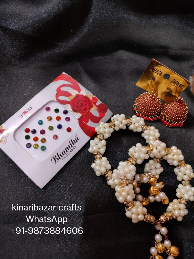 May include: A collection of Indian jewellery and accessories. The image shows a box of colourful bindi stickers, gold-toned earrings with red detailing, and a beaded necklace with pearl-like beads and gold accents. The items are displayed against a dark background.