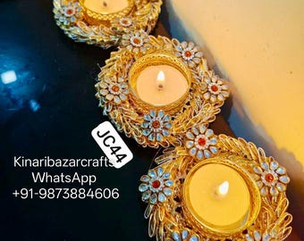May include: Three gold-colored candle holders with lit candles. Each holder is adorned with floral and leaf-like embellishments, and small red and clear gems. The text "JC44" is visible.