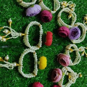 May include: A set of white pearl bracelets with gold accents and tassels. Each bracelet is adorned with a colorful fabric flower.