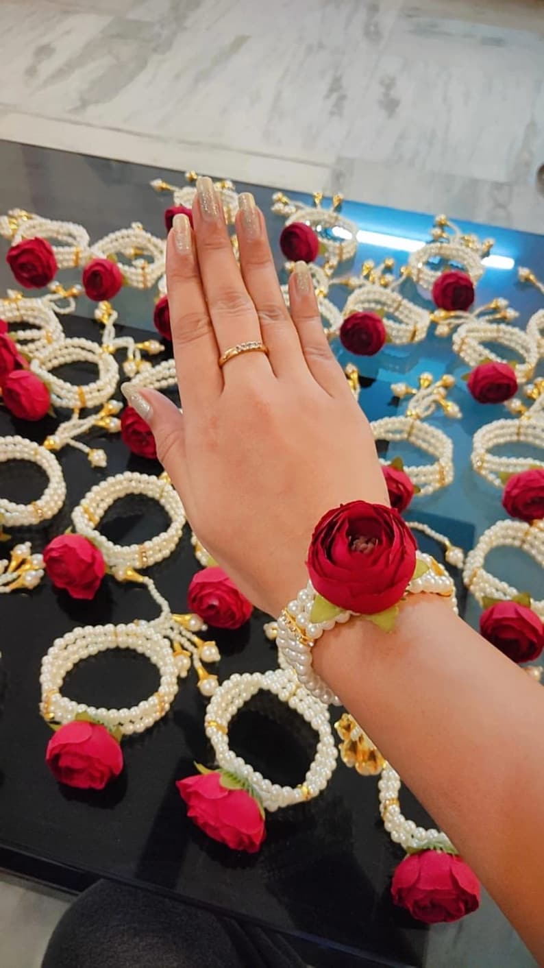 May include: A white pearl bracelet with a red rose attached. The bracelet is on a person's wrist. There are many similar bracelets in the background.