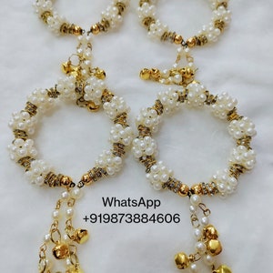 May include: Four white pearl bracelets with gold detailing and hanging gold bells. The bracelets are arranged in pairs, with the text "WhatsApp +919873884606" visible in the image.