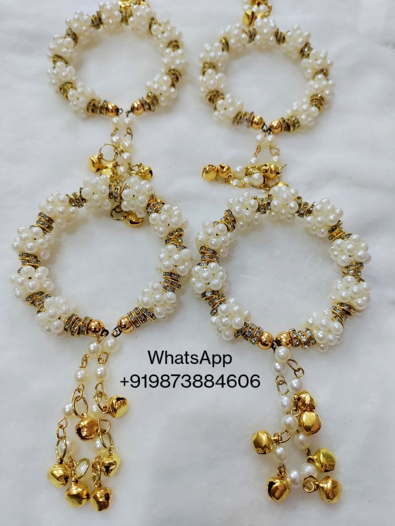 May include: Four white pearl bracelets with gold detailing and hanging gold bells. The bracelets are arranged in pairs, with the text "WhatsApp +919873884606" visible in the image.
