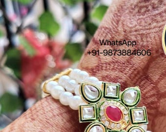 May include: A white pearl bracelet with a gold and green floral design featuring a red gemstone in the center. The bracelet has a WhatsApp contact number: +91-9873884606.