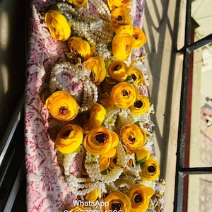 May include: A collection of yellow fabric flower embellishments with pearl bead strings. The flowers are arranged in a pile on a patterned fabric surface. The text "WhatsApp +91-9873884606" is visible at the bottom of the image.