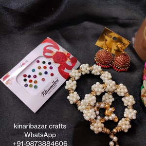May include: A collection of Indian jewellery and accessories against a black backdrop. The items include a white pearl necklace, red and gold earrings, and a box of colourful bindi stickers. The jewellery is displayed with a focus on the details and textures.