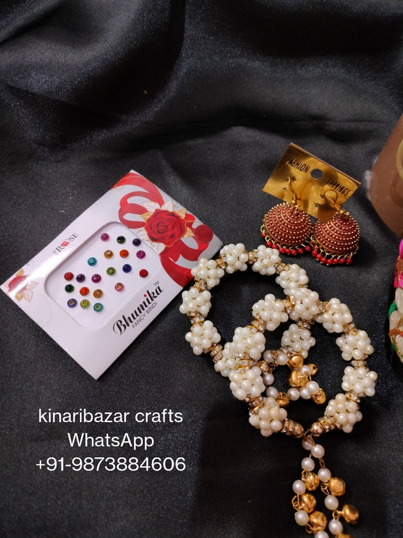 May include: A collection of Indian jewellery and accessories against a black backdrop. The items include a white pearl necklace, red and gold earrings, and a box of colourful bindi stickers. The jewellery is displayed with a focus on the details and textures.