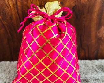 May include: A vibrant pink satin gift bag with a gold diamond pattern. The bag is tied with a pink drawstring and has a gold satin top. The background is a wooden surface.