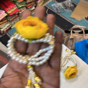 May include: A white pearl bracelet with a yellow flower charm and a gold tassel. The bracelet is on a hand.