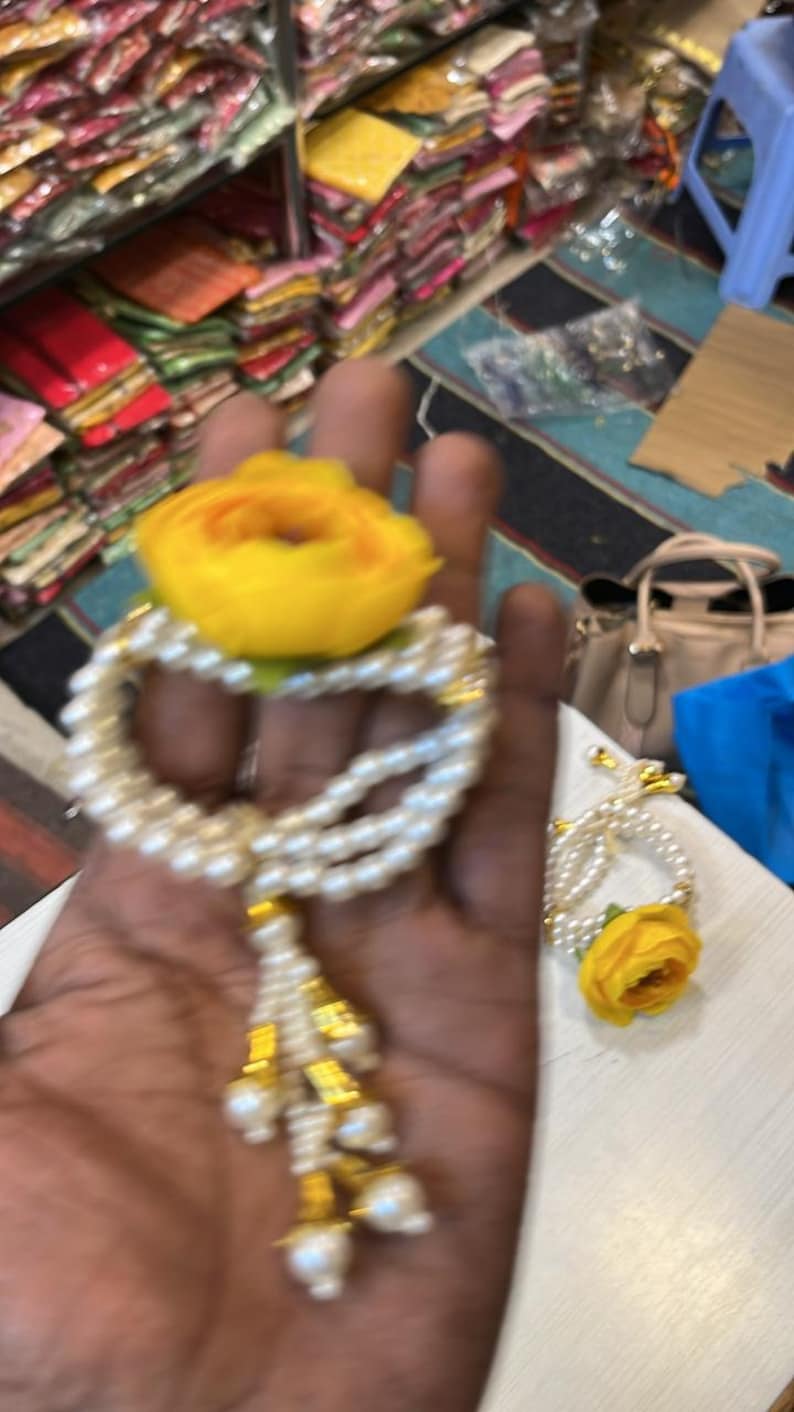 May include: A white pearl bracelet with a yellow flower charm and a gold tassel. The bracelet is on a hand.