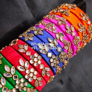May include: A stack of colorful bangles in shades of green, red, blue, pink, and orange. Each bangle is adorned with gold-colored embellishments and clear, sparkling stones. The text "WhatsApp +91-9873884606" is visible.
