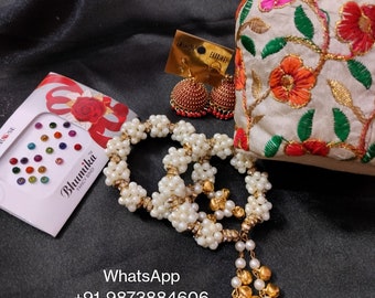 May include: An assortment of Indian jewelry and accessories on a black satin surface. Items include a beaded necklace, earrings, a small embroidered bag, and a box of colorful bindi stickers. The bag is cream-colored with floral embroidery.