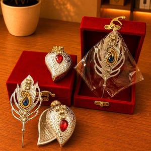 Puede incluir: Una colección de objetos decorativos de metal plateado que incluye dos broches en forma de pluma con cristales azules y transparentes, y dos recipientes en forma de lágrima con cristales rojos, presentados en cajas de terciopelo rojo con cierres dorados.