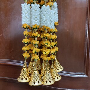 May include: A garland of yellow and white artificial flowers with gold bells hanging from the bottom.