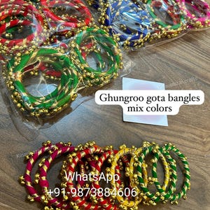 Puede incluir: Surtido de pulseras coloridas con ribetes dorados y pequeñas campanas. Las pulseras están hechas de tela y están dispuestas en una pila. El texto "Ghungroo gota bangles mix colors" y "WhatsApp +91-9873884606" es visible en la imagen.