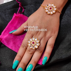 Può includere: Un set di anello e braccialetto in oro con un design floreale. L'anello e il braccialetto presentano una pietra rossa centrale circondata da pietre chiare. L'anello è su un dito con smalto per unghie turchese.