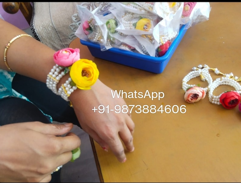 May include: A set of pearl bracelets with colorful fabric flowers. The bracelets are white with gold accents and feature pink, yellow, and red flowers. The text "WhatsApp +91-9873884606" is visible in the image.