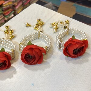 May include: Three white pearl bracelets with red flower accents and gold tassels. The bracelets are arranged on a white surface.