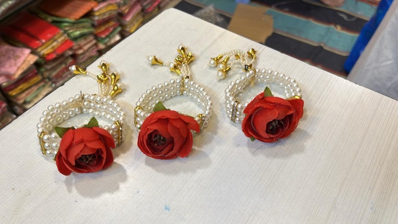 May include: Three white pearl bracelets with red flower accents and gold tassels. The bracelets are arranged on a white surface.