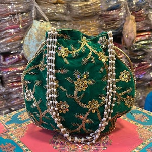 Peut inclure: Un sac à cordon en velours vert avec des broderies dorées et une bandoulière en chaîne de perles. Le sac est posé sur une boîte à motifs rose et bleu.
