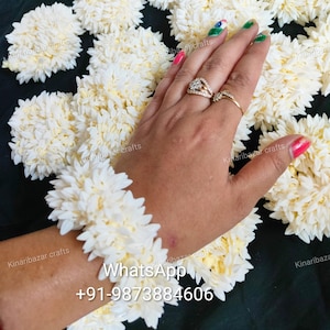 May include: A white flower garland bracelet made of small white flowers. The bracelet is on a person's wrist. The image also shows a variety of other white flower garlands.