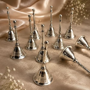 May include: A collection of ten silver-coloured metal hand bells, each featuring a long, slender handle and a flared base with horizontal ring detailing. The bells are arranged in a group, with some standing upright and others lying on their sides.