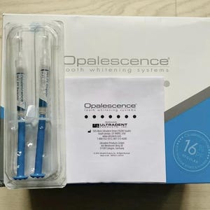 Peut inclure: Un coffret de systèmes de blanchiment dentaire Opalescence. La boîte est blanche avec des accents bleus et contient deux seringues avec des pistons bleus. Une petite brochure blanche et un plateau en plastique transparent sont également visibles.