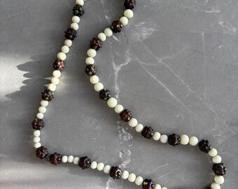 Puede incluir: Un collar con cuentas crema y marrón oscuro alternadas. Las cuentas marrón oscuro tienen un diseño esférico texturizado, con toques de verde y amarillo. El collar se muestra sobre una superficie gris.