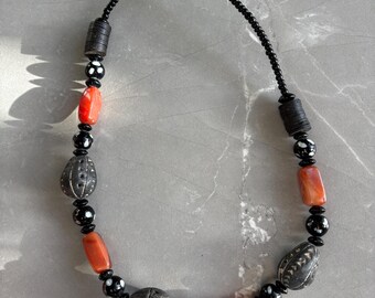 Puede incluir: Un collar artesanal con una mezcla de cuentas negras, plateadas y coral. El collar incluye cuentas plateadas texturizadas, pequeñas cuentas negras y cuentas de color coral vibrante, ensartadas en un cordón negro. El collar se exhibe sobre una superficie gris.