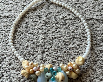 Puede incluir: Un collar de cuentas blancas con un grupo decorativo. El grupo incluye cuentas en forma de lágrima azul claro, pequeñas perlas y detalles en forma de concha en tonos beige y amarillo. El collar está sobre una superficie gris texturizada.