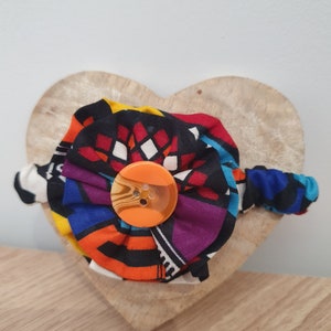 May include: A colourful fabric headband with a large orange button in the centre. The headband is displayed on a wooden heart-shaped surface.