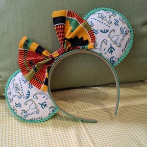 May include: A pair of green and white mouse ears with a yellow, red and black bow. The ears are decorated with a white background and a pattern of lions and tribal symbols.