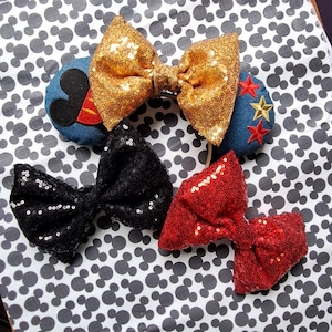 May include: Three pairs of sequined bow headbands. The first headband has a gold bow, the second headband has a black bow, and the third headband has a red bow.