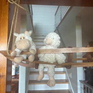 May include: Two white plush sheep toys are sitting on a wooden swing. The sheep are facing forward and have fluffy white fur. The swing is made of wood and has a rope attached to it.