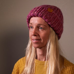 Könnte beinhalten: Eine Frau mit langen blonden Haaren trägt eine rosa gestrickte Mütze mit einem braunen Etikett. Sie trägt einen gelben Pullover.
