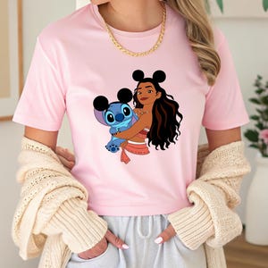May include: A light pink t-shirt featuring a graphic of a cartoon character holding another cartoon character. The graphic includes a woman with long dark hair and a blue alien-like creature. The woman is wearing a red and white outfit.