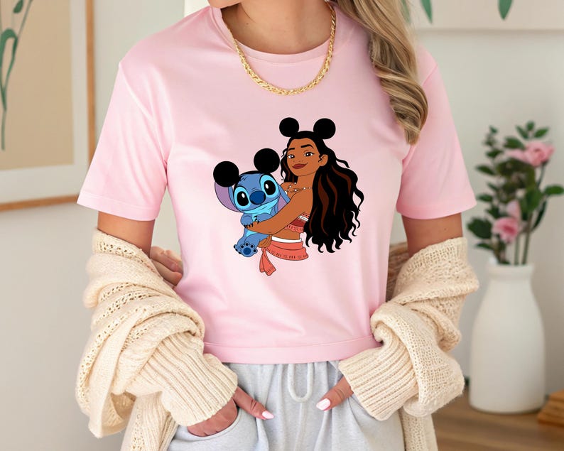 May include: A light pink t-shirt featuring a graphic of a cartoon character holding another cartoon character. The graphic includes a woman with long dark hair and a blue alien-like creature. The woman is wearing a red and white outfit.