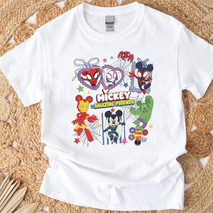 May include: White t-shirt featuring a colorful graphic of Mickey Mouse and other Marvel characters, including Spider-Man, Iron Man, and Hulk. The text "Mickey Amazing Friends" is also displayed. The design is surrounded by stars and webs.