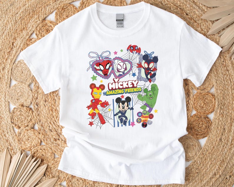 May include: White t-shirt featuring a colorful graphic of Mickey Mouse and other Marvel characters, including Spider-Man, Iron Man, and Hulk. The text "Mickey Amazing Friends" is also displayed. The design is surrounded by stars and webs.