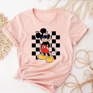 May include: Pink t-shirt with a black and white checkered pattern and a cartoon image of Mickey Mouse wearing sunglasses. Mickey Mouse is wearing red shorts and black shoes.