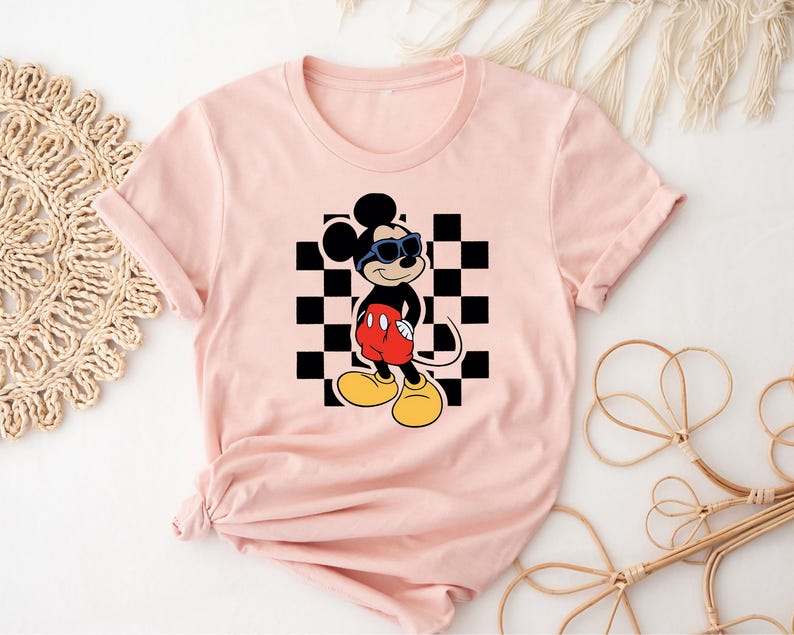 May include: Pink t-shirt with a black and white checkered pattern and a cartoon image of Mickey Mouse wearing sunglasses. Mickey Mouse is wearing red shorts and black shoes.