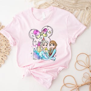 May include: Pink t-shirt with a watercolor illustration of Elsa and Anna from Frozen standing in front of a castle with a pink bow on top. The illustration is surrounded by stars and sparkles.