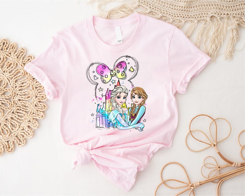 May include: Pink t-shirt with a watercolor illustration of Elsa and Anna from Frozen standing in front of a castle with a pink bow on top. The illustration is surrounded by stars and sparkles.