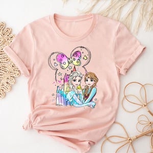 May include: A pink t-shirt with a graphic of Elsa and Anna from Frozen standing in front of a castle with a Mickey Mouse bow on top. The graphic is in a cartoon style and features bright colors.