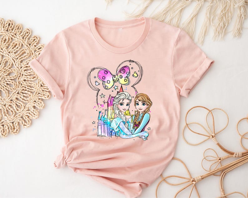 May include: A pink t-shirt with a graphic of Elsa and Anna from Frozen standing in front of a castle with a Mickey Mouse bow on top. The graphic is in a cartoon style and features bright colors.